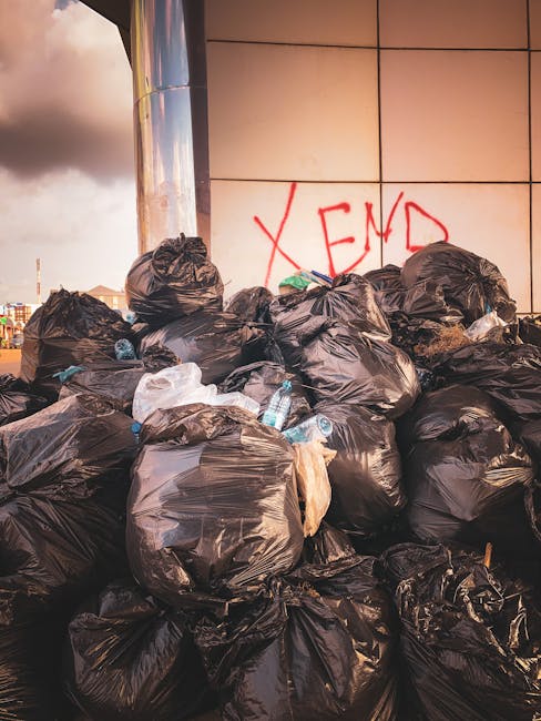A large pile of black plastic rubbish bags, many tied at the top, are stacked against a modern building with beige tiled walls. Some bags are torn or bulging, revealing waste materials such as plastic bottles and paper packaging, which are also scattered around the bags on the ground. The bags are situated outdoors on a paved surface, possibly on a street or parking area, with an overcast sky visible in the background that casts a dull, diffuse light over the scene. On the wall behind the bags, there is red graffiti spelling 'XEND.' The image appears to depict an instance of improper or unmanaged rubbish accumulation, which companies like Rubbish Removal Catford might handle through their private waste collection services, emphasizing the importance of proper rubbish removal and disposal methods.