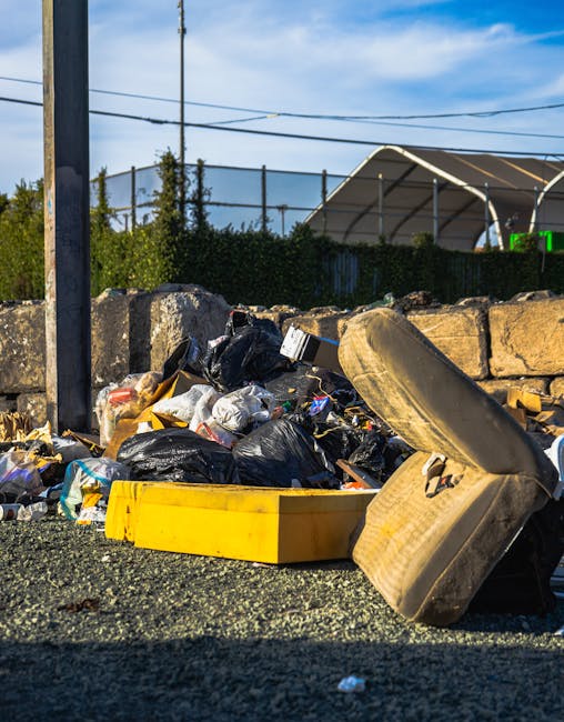A large pile of black plastic rubbish bags, many tied at the top, are stacked against a modern building with beige tiled walls. Some bags are torn or bulging, revealing waste materials such as plastic bottles and paper packaging, which are also scattered around the bags on the ground. The bags are situated outdoors on a paved surface, possibly on a street or parking area, with an overcast sky visible in the background that casts a dull, diffuse light over the scene. On the wall behind the bags, there is red graffiti spelling 'XEND.' The image appears to depict an instance of improper or unmanaged rubbish accumulation, which companies like Rubbish Removal Catford might handle through their private waste collection services, emphasizing the importance of proper rubbish removal and disposal methods.