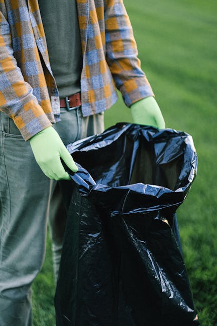 A person wearing a yellow and grey checkered shirt over a green top, with grey trousers and a brown belt, is seen from the waist down, holding open a large black rubbish bag lined with silver foil. The individual is wearing light green gloves and standing outdoors on a grassy area. The black rubbish bag appears partially filled and is positioned upright, ready for waste disposal. The environment suggests an outdoor setting suitable for rubbish removal or cleaning activities, and the image reflects private waste handling efforts akin to independent rubbish collection services often provided by companies like Rubbish Removal Catford. The natural lighting highlights the texture of the rubbish bag's shiny surface and the grass in the background, emphasizing the scene's focus on waste clearance and rubbish disposal processes.