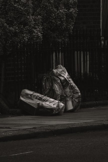 On a quiet street corner, three large black plastic garbage bags filled with waste are placed directly on the pavement near a dark metal fence and a hedge of leafy trees. The bags are tightly knotted at the top, appearing to contain household rubbish or yard debris, with some areas showing reflective or glossy surfaces that catch minimal light in the subdued environment. Behind the fence, there is a partial view of a building structure, indicating an urban or suburban setting. The scene suggests an instance of private waste disposal, potentially associated with non-regular rubbish collection, emphasizing the need for professional rubbish removal services in Catford, such as those offered by Rubbish Removal Catford. The overall composition is neutral and factual, with an emphasis on the straightforward visual of waste awaiting collection, reflective of typical private waste handling methods.