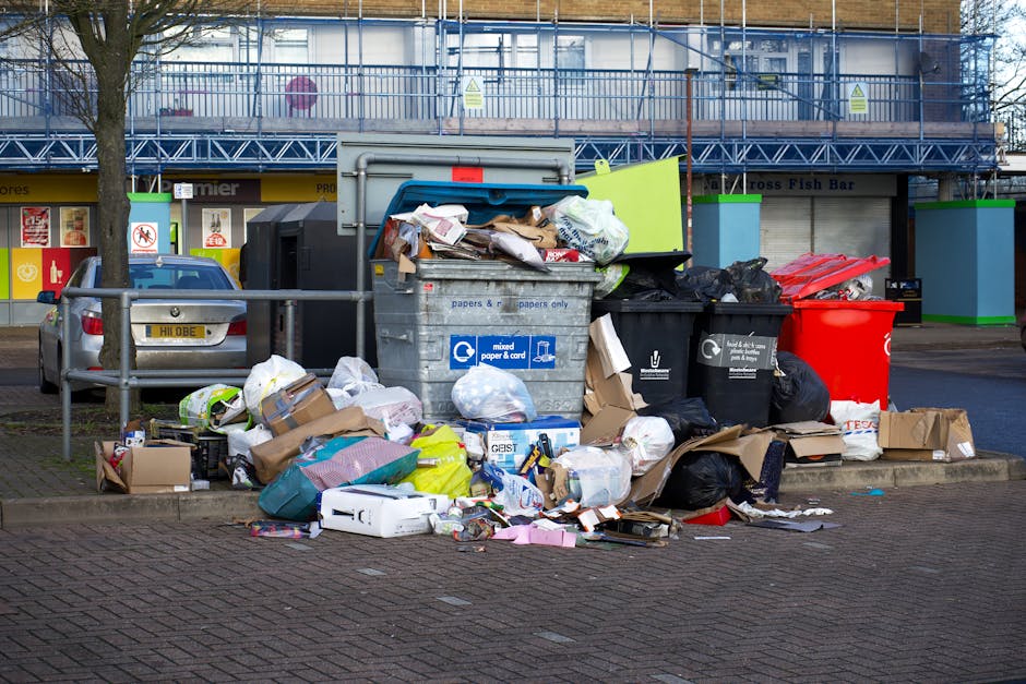 A large collection of mixed waste and rubbish bags, including cardboard boxes, plastic packaging, paper, and black rubbish bags, is piled next to a grey metal recycling bin marked for paper and cardboard, situated on a paved area outside a commercial building. The rubbish spills over onto the sidewalk, with some items partially on the curb and others leaning against the bin. Behind the pile, there are two parked cars, one grey and the other blue, with a low metal railing separating the parked vehicles from the rubbish area. The background features a storefront with signage and a building under construction or renovation, indicated by scaffolding and blue safety netting. The scene is lit by natural daylight, and the environment appears to be an urban commercial or retail area, where independent waste disposal or inappropriate rubbish accumulation is evident, highlighting the importance of proper rubbish management and adherence to local waste collection regulations, as managed by services like Rubbish Removal Catford.