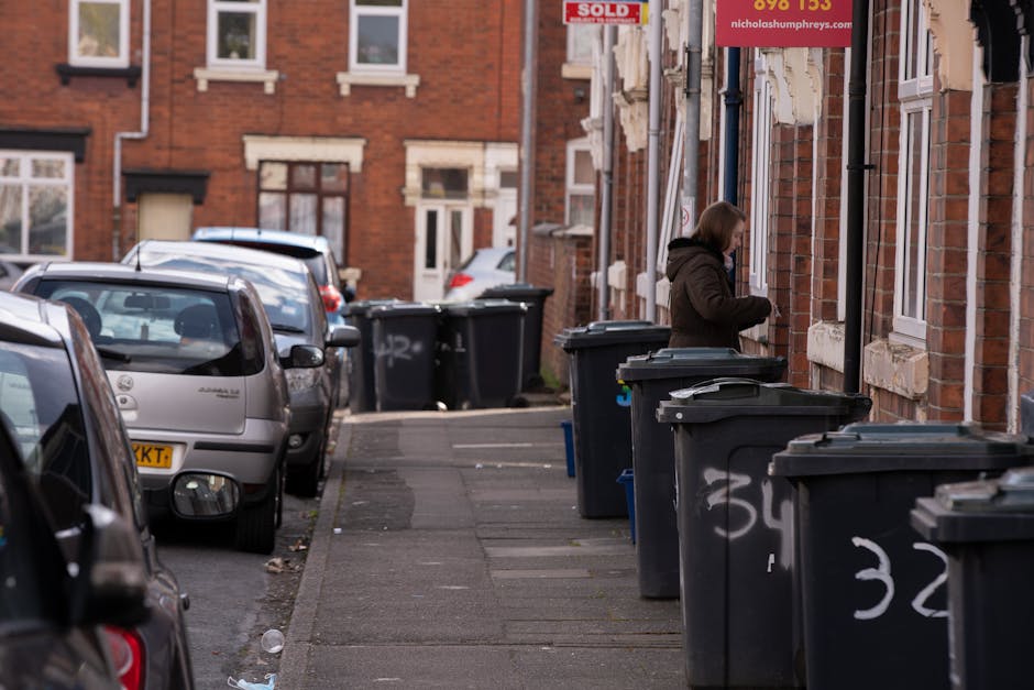 A residential street scene showing a woman standing beside a series of black wheelie bins lined up along the pavement in front of a red-brick building. The bins are made of plastic with lids and vary slightly in size, positioned side by side close to the building's wall. The woman, dressed in a dark brown coat, appears to be handling or opening one of the bins. To the left, several parked cars occupy the street, including a silver hatchback and a darker vehicle, with some visible front and rear ends. The background features a row of similar brick houses with white-framed windows and small front doors, with some windows partially obscured by the bins. The pavement surface is made of concrete slabs, some showing minor wear, and a few small pieces of litter are scattered near the parked cars. Bright, natural daylight illuminates the scene, highlighting the urban environment typical of a residential area where private waste collection through bin storage outside is common, supporting alternative approaches to scheduled rubbish removal services by local councils. Rubbish Removal Catford’s service focus on managing such waste materials aligns with the scene depicted, emphasizing on-site or independent waste disposal options. The image overall reflects a typical street setup for rubbish management, including allocated bin storage, and highlights the importance of adhering to local rubbish disposal regulations. The overall composition and detail capture both environmental context and material textures clearly, suitable for accessibility and SEO relevance in waste management content.