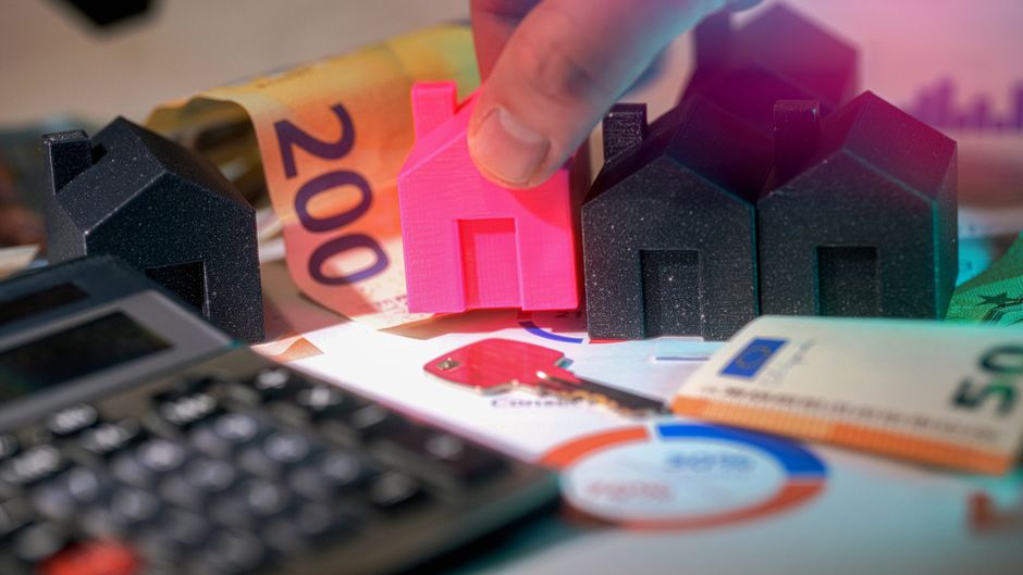 A close-up image features a person's finger pressing a small, bright pink model house among other miniature black houses positioned on a cluttered surface. Surrounding the models are various financial documents, including printed charts and colorful paper cutouts, alongside a calculator and euro banknotes in denominations of 50 and 200. The scene appears to represent concepts of property, housing valuation, or real estate costs, with the models symbolizing residential properties. The background is slightly blurred, emphasizing the models and financial elements in the foreground, suggesting a context of property valuation or private housing expenses, which aligns with themes related to house clearance or independent property management services provided by Rubbish Removal Catford. The lighting is even, with no harsh shadows, creating a neutral and professional atmosphere suitable for informational or service-related content about property and waste disposal costs.