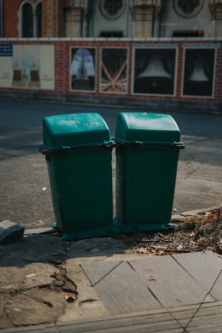 Three large wheeled rubbish bins are positioned in a row on the edge of a pavement, each made of durable plastic with a textured finish. The left bin is black with a closed lid and stands upright, while the middle bin is blue with a partially open lid, slightly taller with more prominent ridges on its surface. The right bin is green, also with a closed lid, and appears to be slightly bent at the front. All three bins are placed on a curb's concrete edge, with small patches of grass and weeds visible close to their bases. Behind the bins, a dense hedge of leafy green foliage forms the background, with hints of branches and trunks appearing through the dense leaves. The scene is illuminated by natural daylight, providing clear visibility of the colours, textures, and details of the bins and surrounding environment, consistent with a typical residential area that may engage services from Rubbish Removal Catford for waste collection and alternative disposal methods.